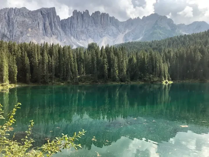 Lago verde smeraldo circondato da abeti, con le guglie delle Dolomiti sullo sfondo